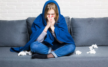 Sick Woman With Rheum And Headache Holding Napkin, Sitting On Sofa With Coveret And Pills At Home