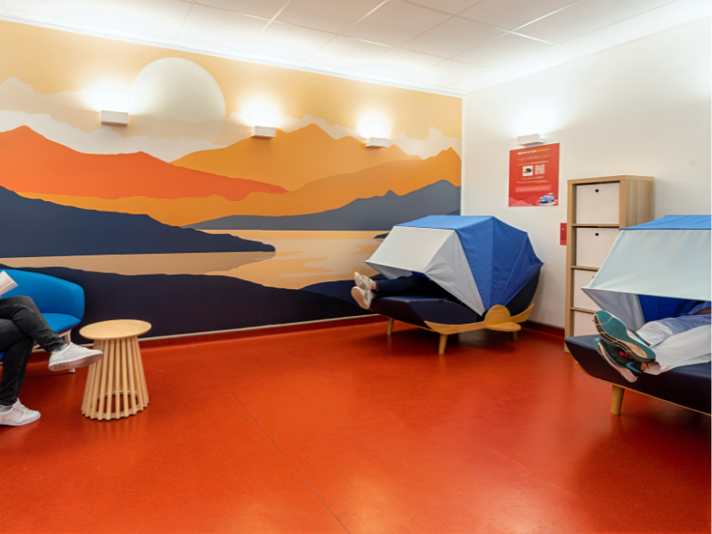 Vue intérieure de la salle de détente « La Parenthèse ». L’espace est chaleureux et apaisant, avec un sol rouge, un grand mur décoré d’une fresque aux teintes orangées représentant un paysage de montagnes au coucher du soleil. Deux cocons de repos inclinés avec coussins bleus sont disposés sur la droite, à côté de casiers de rangement. Un canapé bleu et une petite table en bois clair complètent l’aménagement sur la gauche. L’ambiance lumineuse est douce, avec des appliques murales diffusant une lumière indirecte. 